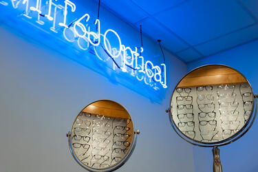 MIT Pptical neon sign above two round desk mirrors showing the reflection of eyeglasses in display racks