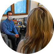 A patient and clinician look toward each other. The patient with blond and brown streaked hair is seen from the shoulders up from the rear. The clinician wears a PPE mask and is seated and visible head to toe sitting in comfortable office chair while facing patient.