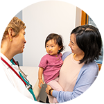 Side view of a pediatrician wearing a white clinical coat talking to a mother holding a toddler-age child who is looking at the pediatrician