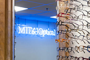 Partial view of eyeglass frames at n optical store, with a neon sign visible in the mirror reading, 'MIT Optical'