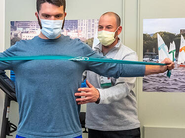 Sports Medicine and Orthopedics clinician standing behind a patient dressed in workout clothing, with arms outstretched holding a physical therapy band. Both people are seen from the waist up, standing, and wearing PPE masks.