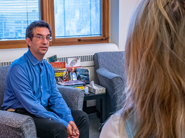 Full frame view of mental health clinician sitting in a comfortable chair and looking forward and to the right of the frame at a student, whose head and shoulders are visible from behind on the right side of the sr