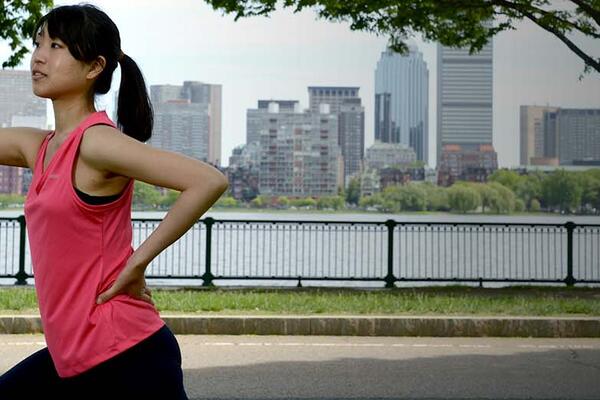 Waist up view of woman wearing athletic clothing with one arm against a tree and the other akimbo. A tree, the Charles River and Boston skyline are visible in the background.