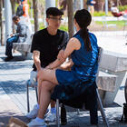 Full view of two MIT students sitting outdoors on a bench with a grassy area and several other people on benches and chairs are visible in the background. The students look toward each other with one students face visible and the other seen from behind.