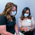 waist up view of a patient and a clinician, both wearing PPE masks. Both look at a clipboard held by the clinician