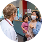 Patient wearing a PPE mask and holding an unmasked toddler, talks with a clinician, masked and seen from the rear, in MIT Medical’s Primary Care waiting area